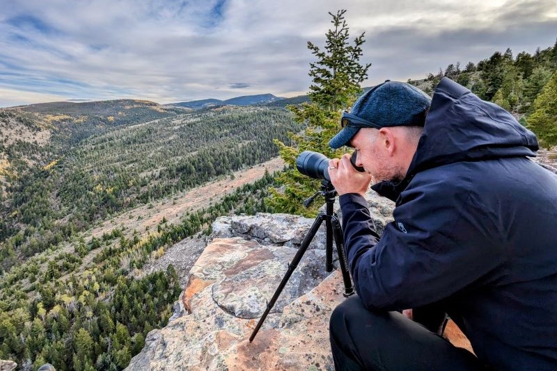 man on hilltop couching to look into camera on a tripod perched on a rock, overlooking forests below